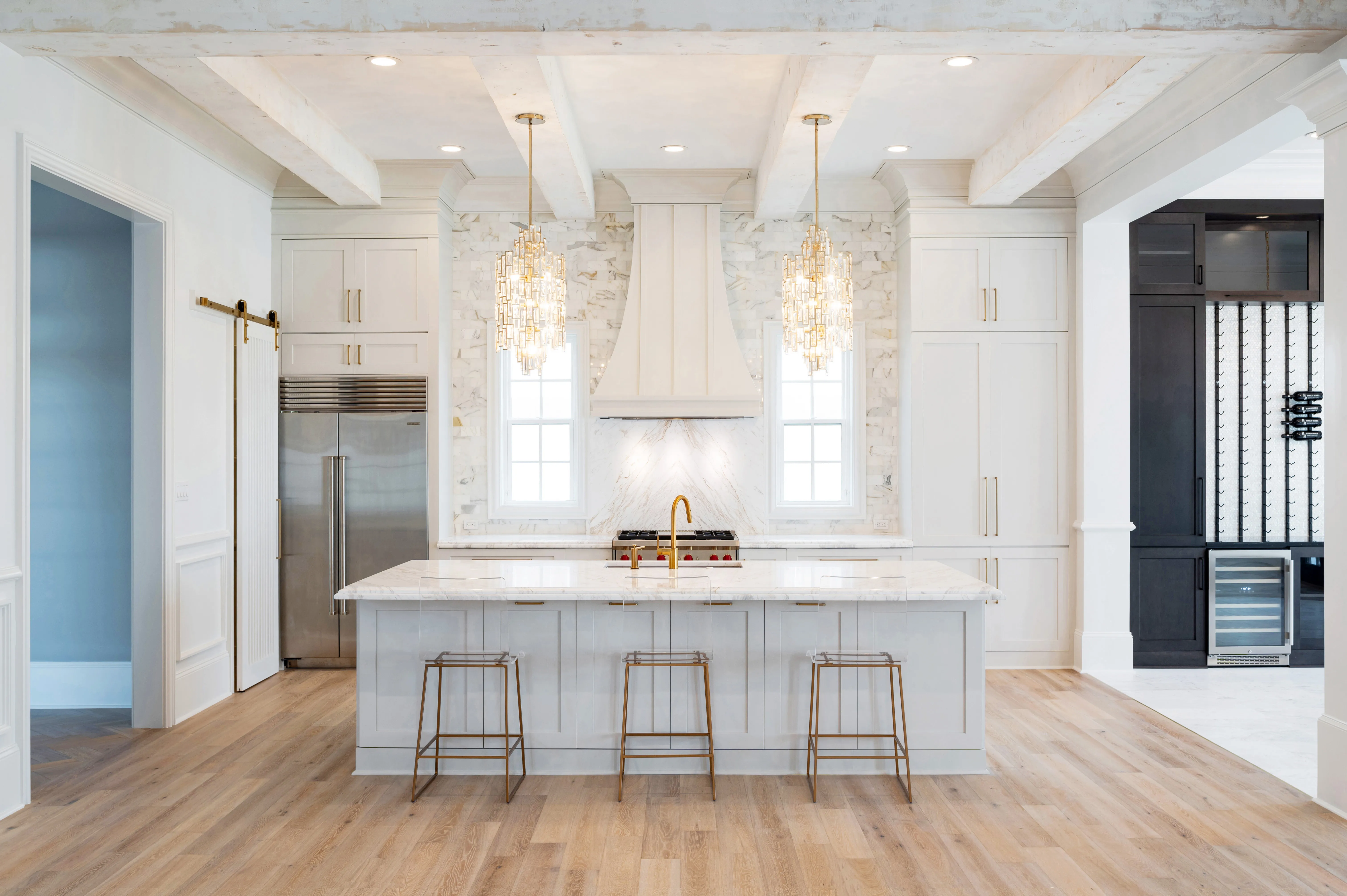 All-white kitchen under painted beams with a marble hood wall and crystal pendants